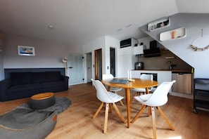 A modern living room with a dark sofa, a round wooden dining table with white chairs, a kitchen with white cabinets, and a staircase leading to another level. - Möwennest by DeJu Norderney (3*** DTV) (Norderney)