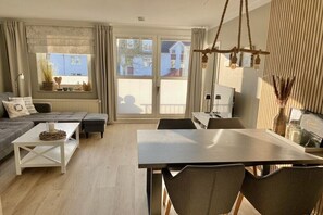 A modern living room with a sofa, a dining table, and chairs. There is a large window with a view of buildings outside. - Large Baltic Sea nest (Boltenhagen)