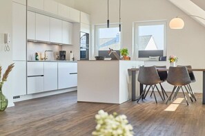 Eine moderne Küche mit weißen Schränken, einem Essbereich mit Tisch und Stühlen sowie einem großen Fenster, das für natürliches Licht sorgt.