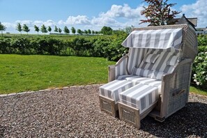 A wicker lounge chair with striped cushions, set on a gravel path with a grassy field and trees in the background.
