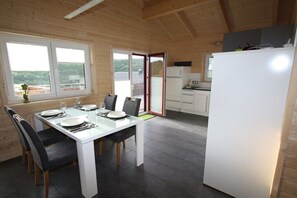 A wooden interior with a dining table set for four, a kitchen with white appliances, and a view through a sliding door. - Holiday home in Rieden with swimming pool (Rieden)