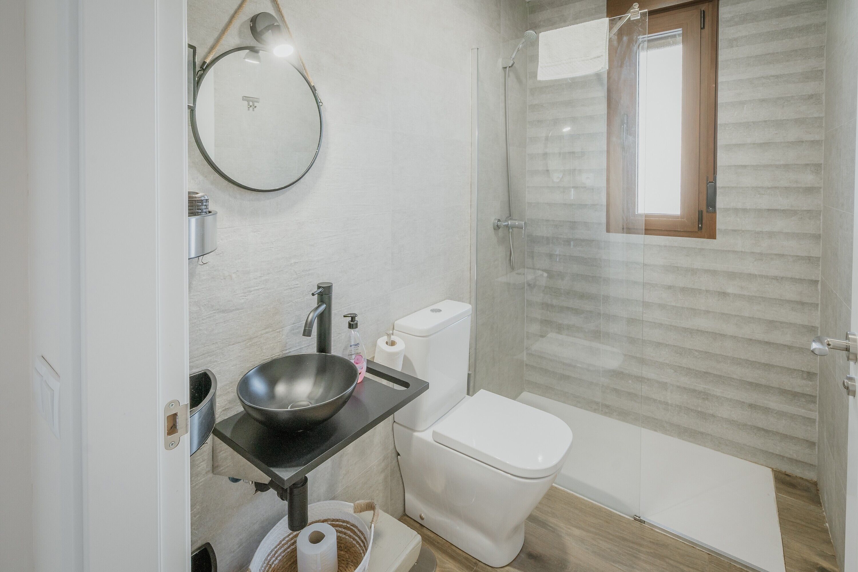 A bathroom with a round mirror, a black sink, a white toilet, and a glass-enclosed shower.