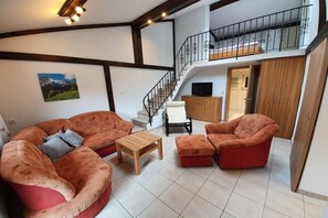 A spacious living area with a curved sofa set, a wooden coffee table, and a TV on a wooden cabinet. A staircase leads to an upper level with a balcony. - Watzmann holiday apartment for couples (Schönau am Königssee)