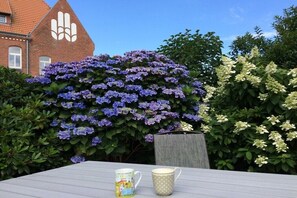 A table with two mugs, a brick building with a distinctive logo, and a bush with purple flowers. - Holiday apartment in Sylt with swimming pool (Sylt)