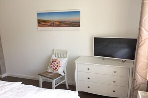 A bedroom with a bed, a chair with a decorative pillow, a white dresser, a television, and a framed picture on the wall. - Holiday apartment in Sylt with swimming pool (Sylt)