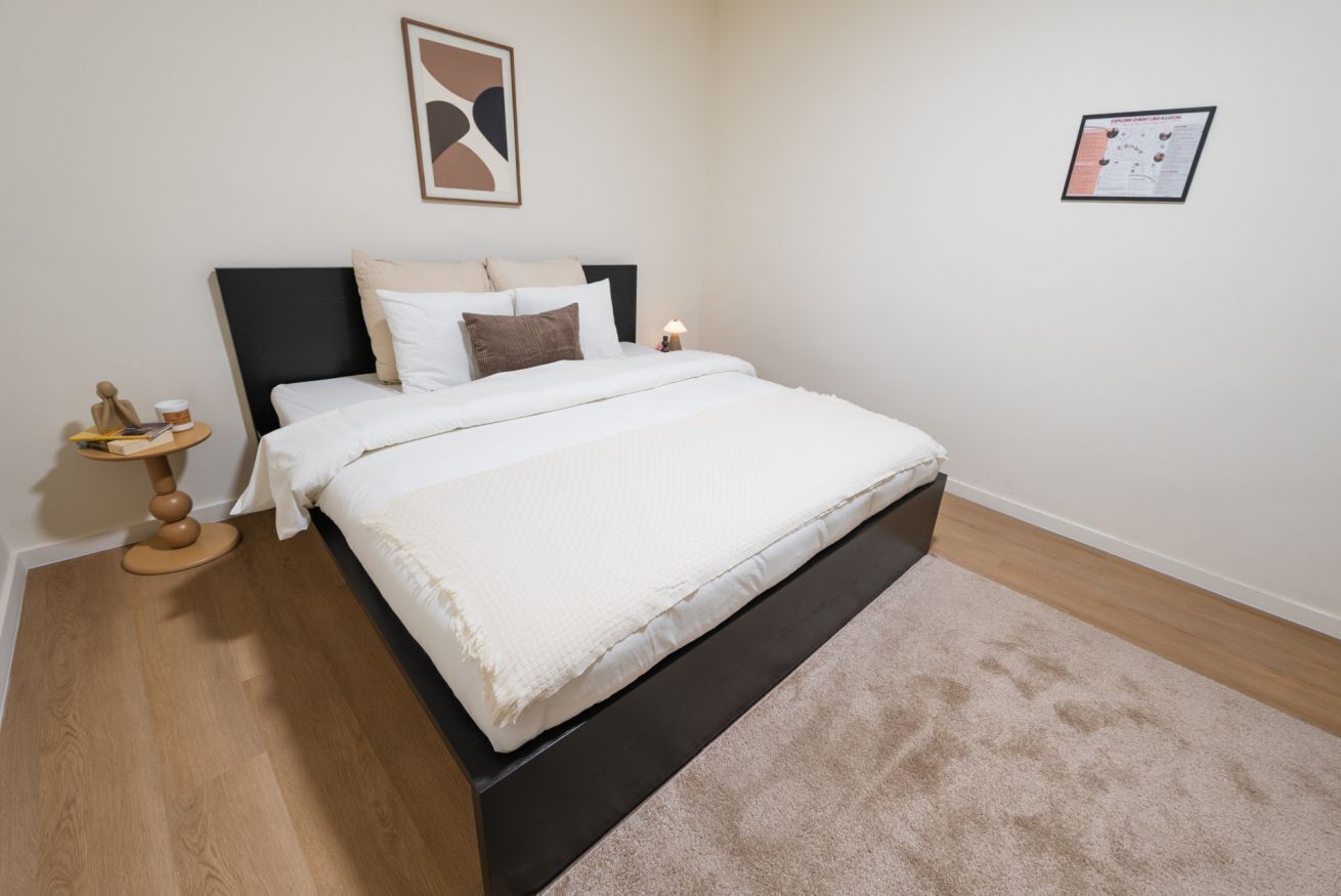 Ein Schlafzimmer mit einem Bett, einem Nachttisch, einer Lampe, einem gerahmten Bild und einem Teppich.