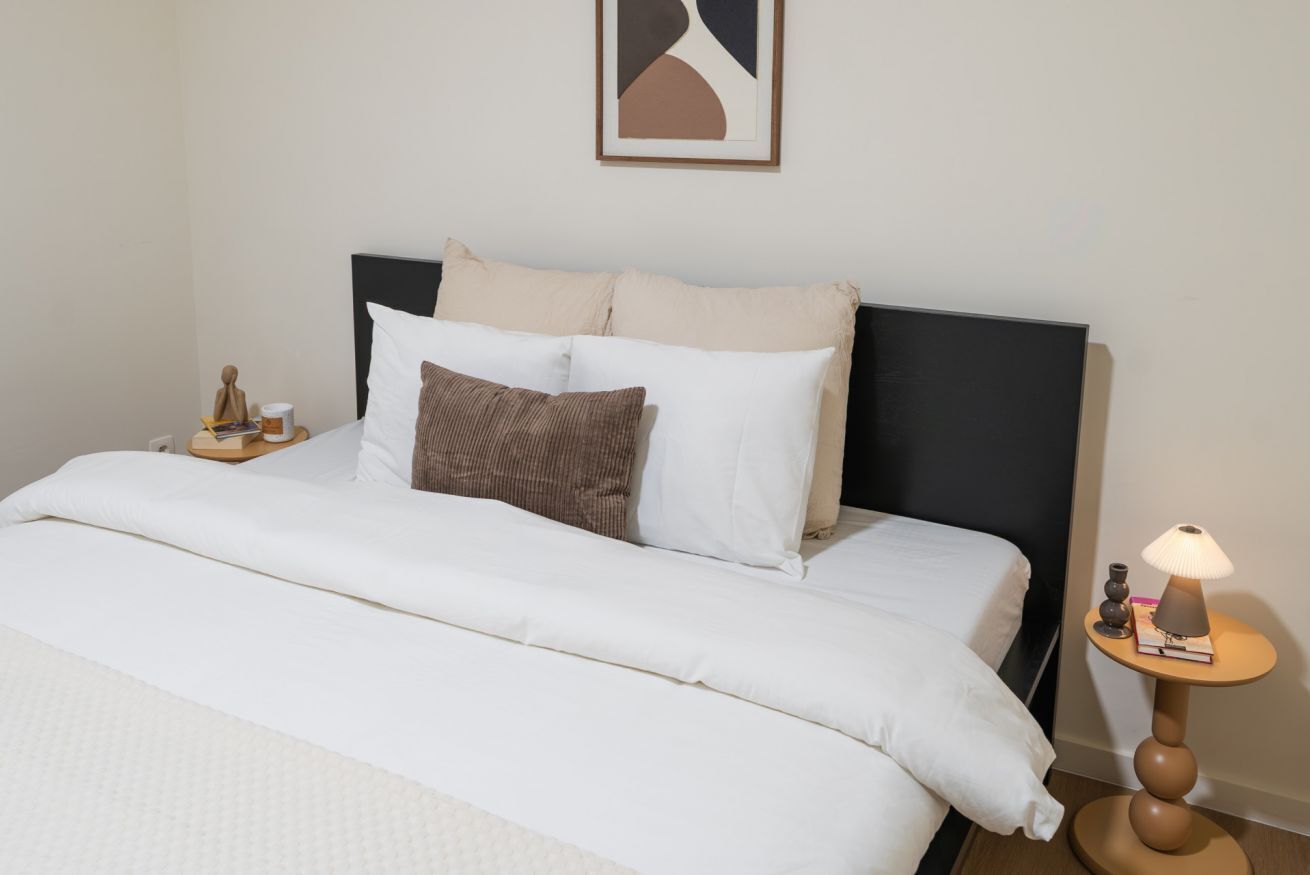 A neatly made bed with white bedding, a black headboard, and a small bedside table with a lamp and a book.