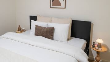 A neatly made bed with white bedding, a black headboard, and a small bedside table with a lamp and a book.