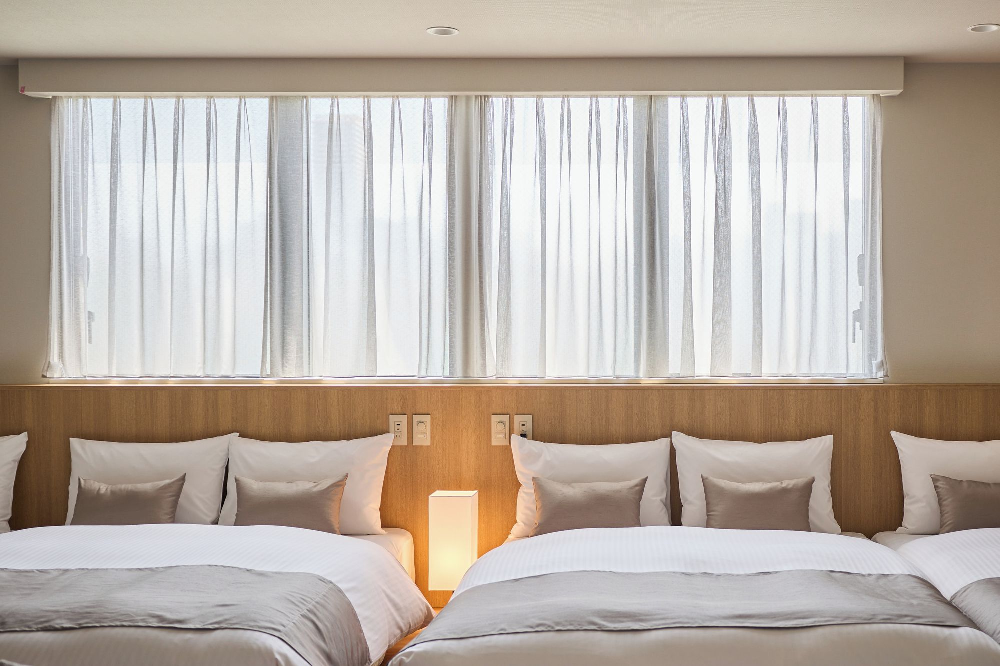 Habitación de hotel con dos camas, un ventanal grande con cortinas translúcidas y un cabecero de madera.