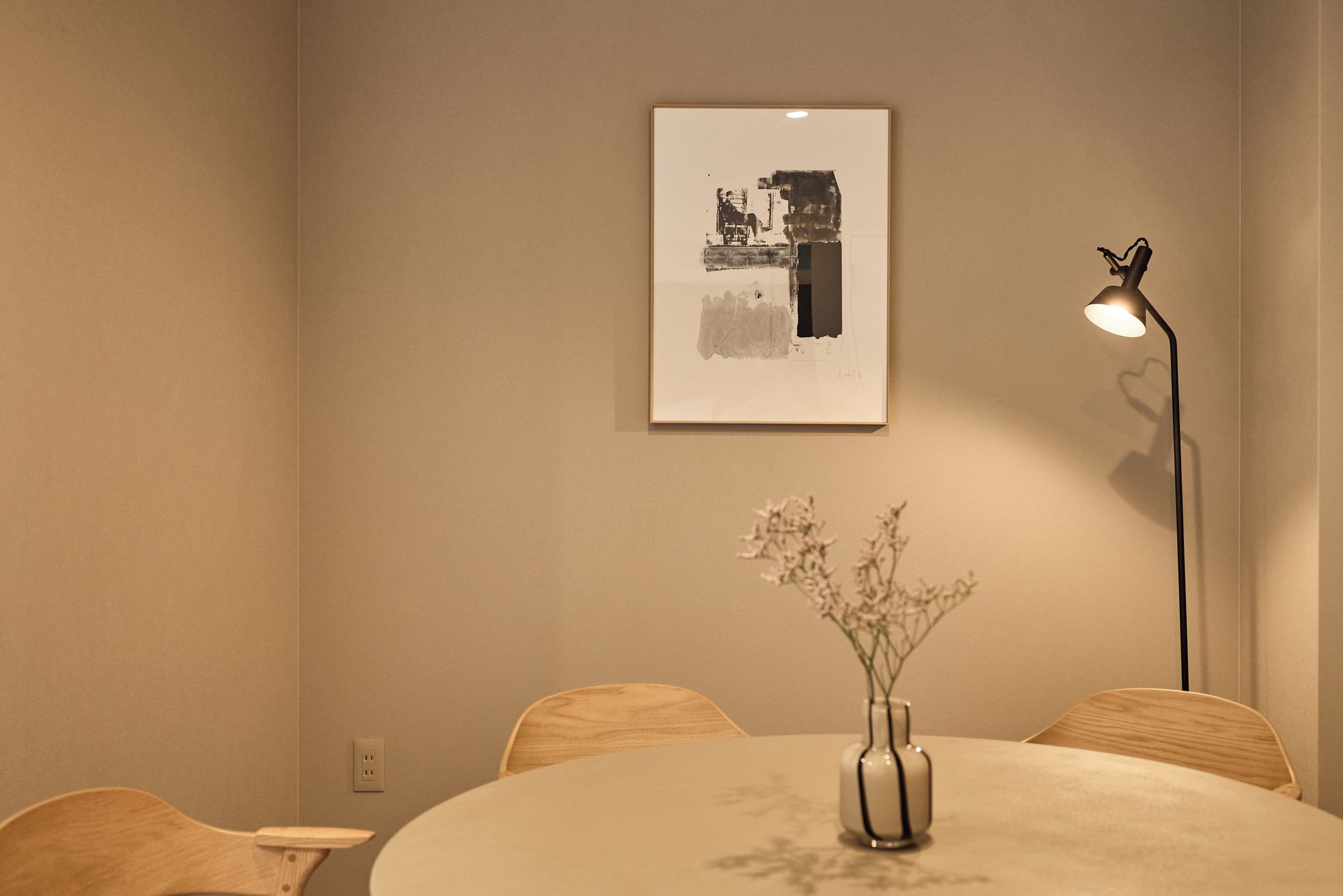 A minimalist room with a round table, a vase with flowers, a framed artwork on the wall, and a floor lamp.