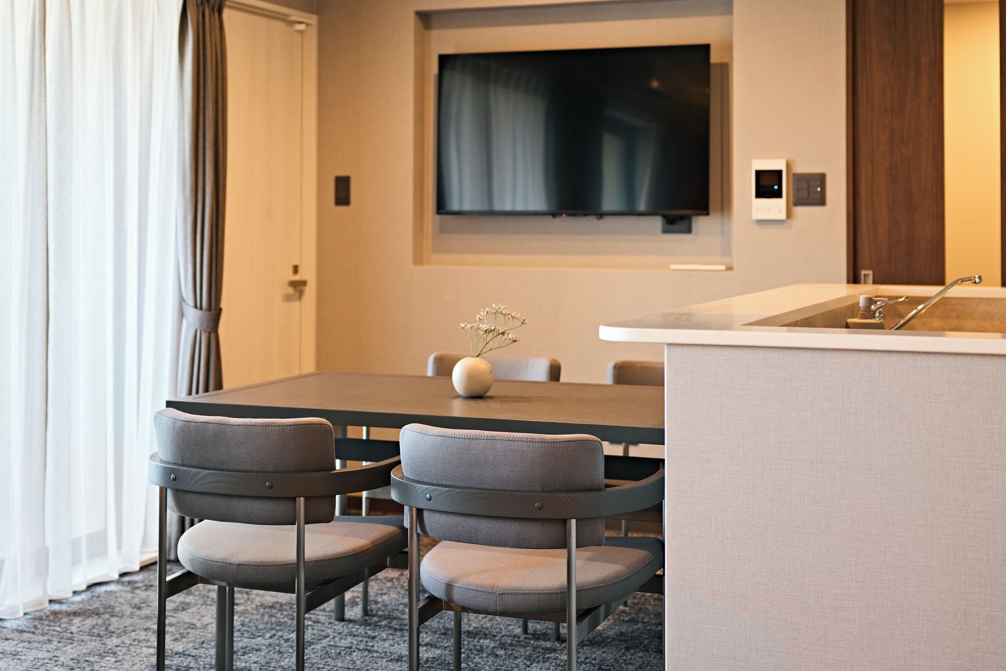 A modern hotel room with a dining area, a flat-screen TV mounted on the wall, a small sink, and a vase with flowers on the table.