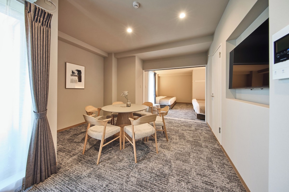 A hotel room with a round dining table, six chairs, a bed, a TV, and a window with curtains.