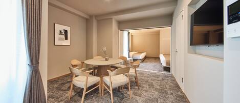 Quarto de hotel com mesa de jantar redonda, seis cadeiras, uma cama, televisão e janela com cortinas.