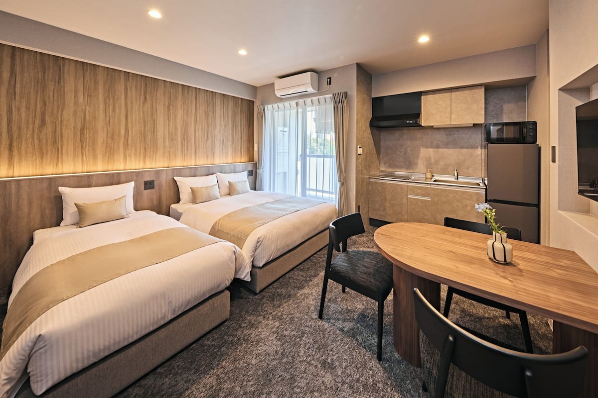 A hotel room with two beds, a dining table, a kitchen area, and a large window with curtains.