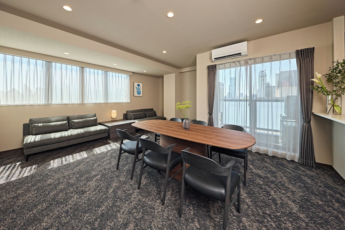 A conference room with a long table, chairs, a sofa, and a large window with curtains.