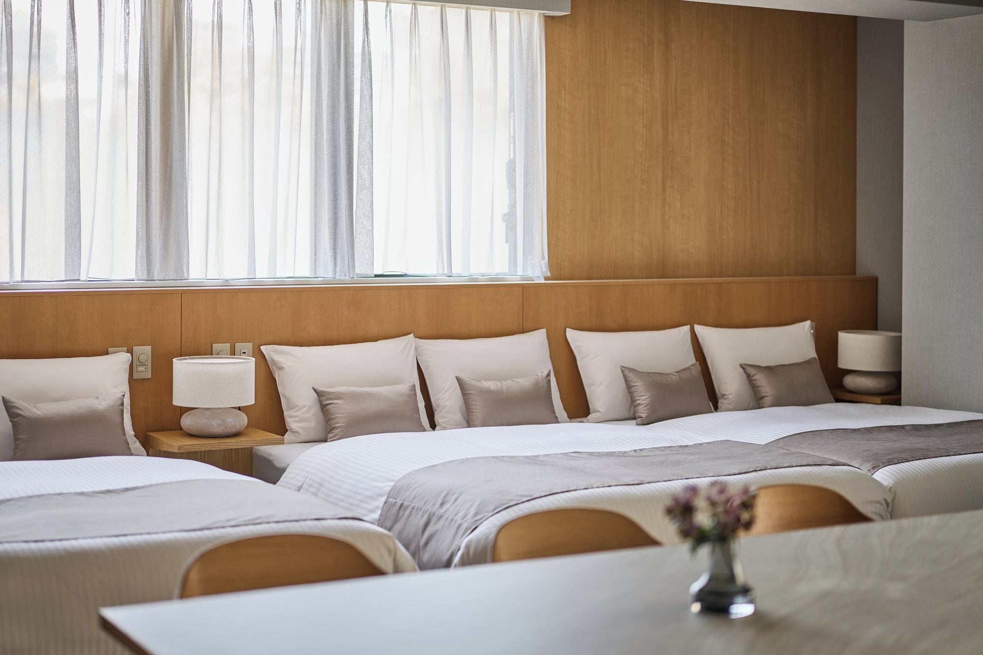 Una camera d'albergo con una testiera in legno, un'ampia finestra con tende leggere, un letto con molti cuscini, lampade sui comodini e un tavolino con un vaso.