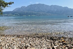 Ein kiesiger Strand mit klarem Wasser, Berge im Hintergrund und ein wolkenloser Himmel.