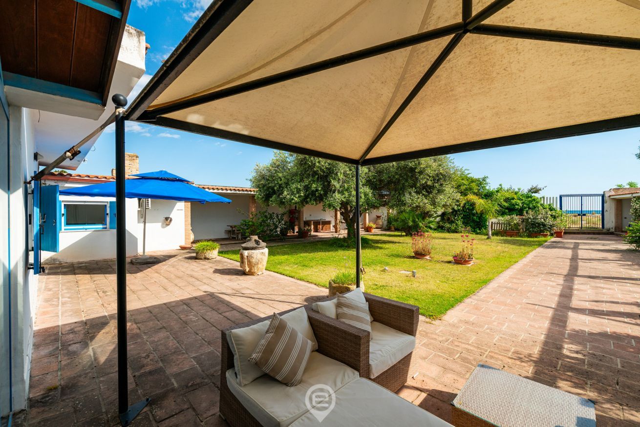 Eine überdachte Terrasse mit einem Korbsessel, einem Tisch und einem blauen Sonnenschirm.