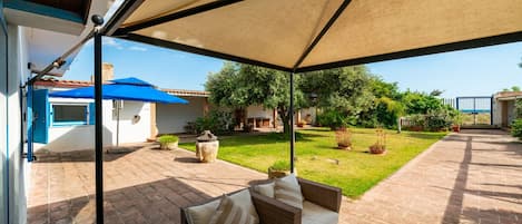 Una veranda riparata con un divano in vimini, un tavolo e un ombrello blu.