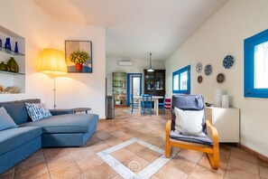 Ein Wohnzimmer mit Sofa, Sessel, Lampe, einer Essecke mit Tisch und Stühlen, einer Küchenzeile und einem Flur, der zu einer Tür führt.