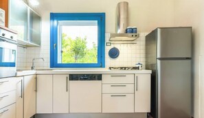 Eine Küche mit weißen Schränken, einem Fenster mit blauem Rahmen, einem Edelstahl-Kühlschrank und einem Geschirrständer.