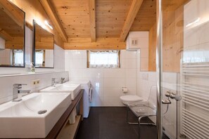 Une salle de bain avec des poutres en bois au plafond, deux lavabos, des toilettes et une douche Ă parois de verre.