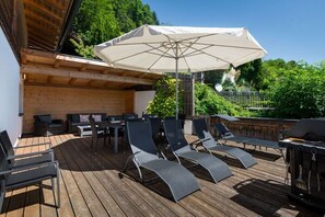 Eine Terrasse im Freien mit Holzboden, einem Tisch mit Stühlen, einem Grill und Liegestühlen unter einem großen Sonnenschirm.