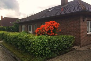 Ein Haus aus Ziegelsteinen mit einem Ziegeldach, einem grünen Heckenstreifen und einem Busch mit roten Blüten.