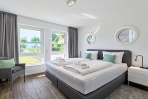Ein modernes Schlafzimmer mit einem großen Bett, zwei Fenstern, einem Sessel und einem Nachttisch.