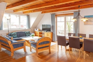 Ein gemütliches Zimmer mit Holzbalkendecke, einer Couch, einem Esstisch mit Stühlen, einem Fernseher und großen Fenstern.