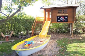 Ein Spielhaus im Piratenthema aus Holz mit Rutsche, Sandkasten und einem Schild mit der Aufschrift 'Piraten-Nest'.