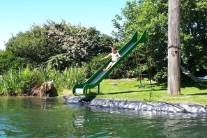 Eine Person auf einer grünen Rutsche über Wasser, umgeben von viel Grün und einer Hängematte.