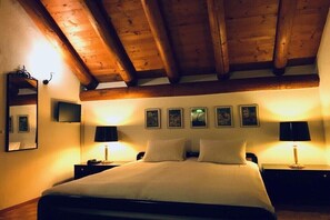 Ein Schlafzimmer mit Holzbalkendecke, einem Bett mit weißen Leinen, zwei Nachttischen mit Lampen und gerahmten Bildern an der Wand.