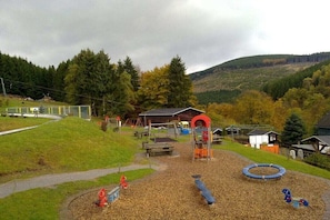 Ein Spielplatz mit Schaukeln, einer Rutsche und einer Kletterkonstruktion, umgeben von Bäumen und einem Hügel.
