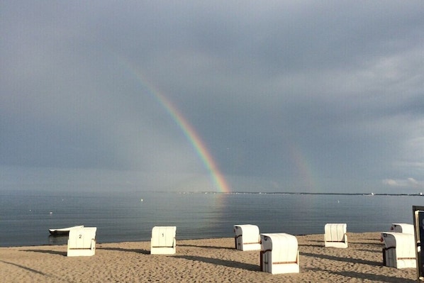 Ein Strand mit weißen Liegestühlen und einem deutlich sichtbaren Regenbogen am Himmel.