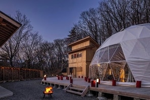 A wooden cabin with a large geodesic dome structure, a fire pit, and a deck with red candles.
