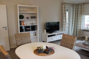 Ein Zimmer mit einem weißen runden Tisch, Korbstühlen, einer weißen Unterhaltungseinheit mit Fernseher und einem Bücherregal mit verschiedenen Gegenständen.