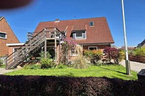 Ein zweigeschossiges Haus mit einer hölzernen Freitreppe, umgeben von einem gepflegten Garten mit verschiedenen Pflanzen und einem Hecken im Vordergrund.