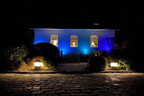 Ein Gebäude mit drei beleuchteten Fenstern bei Nacht, einem Kopfsteinpflasterweg und zwei beleuchteten Laternen im Vordergrund.