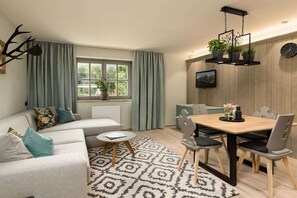 Ein gemütliches Wohnzimmer mit einer Couch, einem Esstisch mit Stühlen, einem gemusterten Teppich, einem Fenster mit Vorhängen und einer an der Wand befestigten Pflanze.