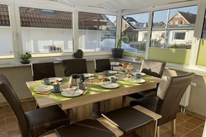 Ein Essbereich mit einem Tisch für sechs Personen, umgeben von Stühlen, großen Fenstern und Blick auf Häuser.