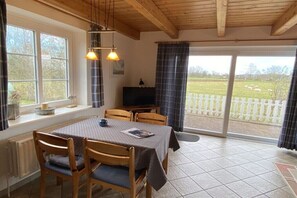 Ein Essbereich mit Tisch, Stühlen, einem Fernseher und Blick auf ein grünes Feld durch eine Schiebetür.