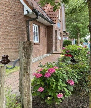 Ein Backsteinhaus mit Ziegeldach, einem Fenster und einem Weg, der zu einem Blumenbeet mit rosa Blüten führt.