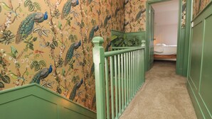 Un couloir orné d’un papier peint à motifs floraux et doté de rampes vertes, qui mène à une chambre.