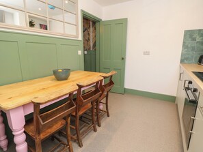 A dining area with a wooden table and chairs, a green door, and a kitchen with a stove. - 8 Manor Villas (Totland Bay)