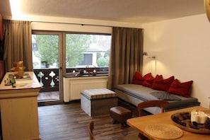 Ein Zimmer mit einer Couch, einem Esstisch, einem Fenster mit Vorhängen, einer Heizung und Blick auf Bäume und Gebäude.
