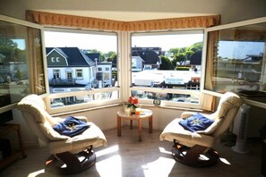 A room with two rocking chairs, a small table with a vase, and a view of buildings through large windows. - Villa by the sea 64-Seaside (Grömitz)