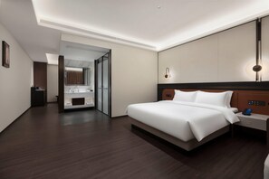 Ein modernes Hotelzimmer mit einem großen Bett, einem Nachttisch mit Telefon und einem durchgegangene Tür einsehbaren Badezimmer.