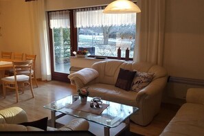 Ein Wohnzimmer mit einer Couch, einem Glastisch, einem Esstisch mit Stühlen und einem Fenster mit Vorhängen.