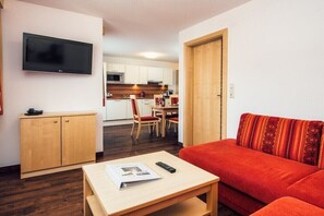 A compact living space with a sofa, a television mounted on the wall, a wooden cabinet, a dining area with chairs, and a kitchen in the background. - Apartment with mountain view (Ladis)
